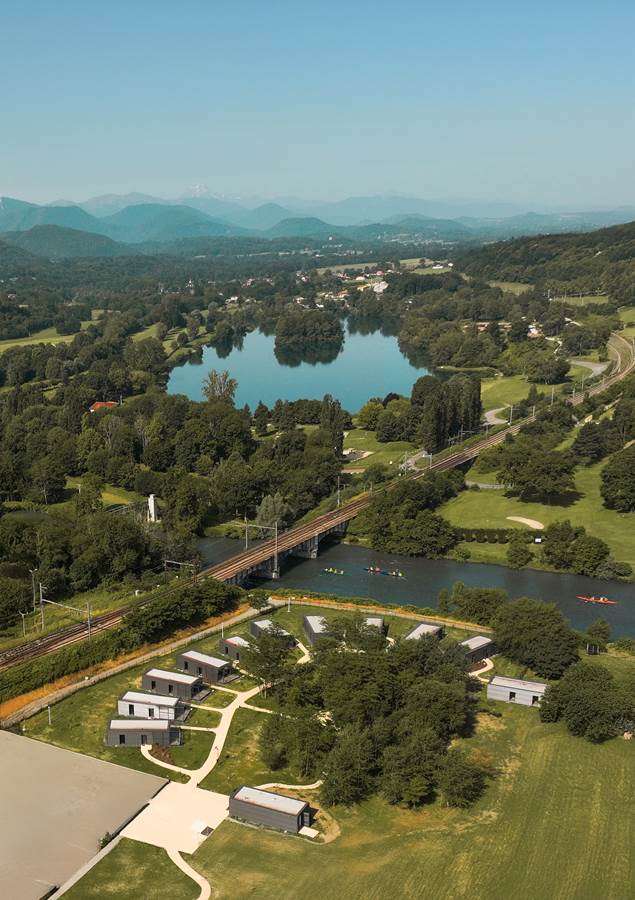 Vue aérienne de l'éco-campus du Comminges à Gourdan-Polignan, douze logements en containers maritimes disposés dans un bois au bord de la Garonne, avec le lac de Montréjeau et les Pyrénées en arrière-plan