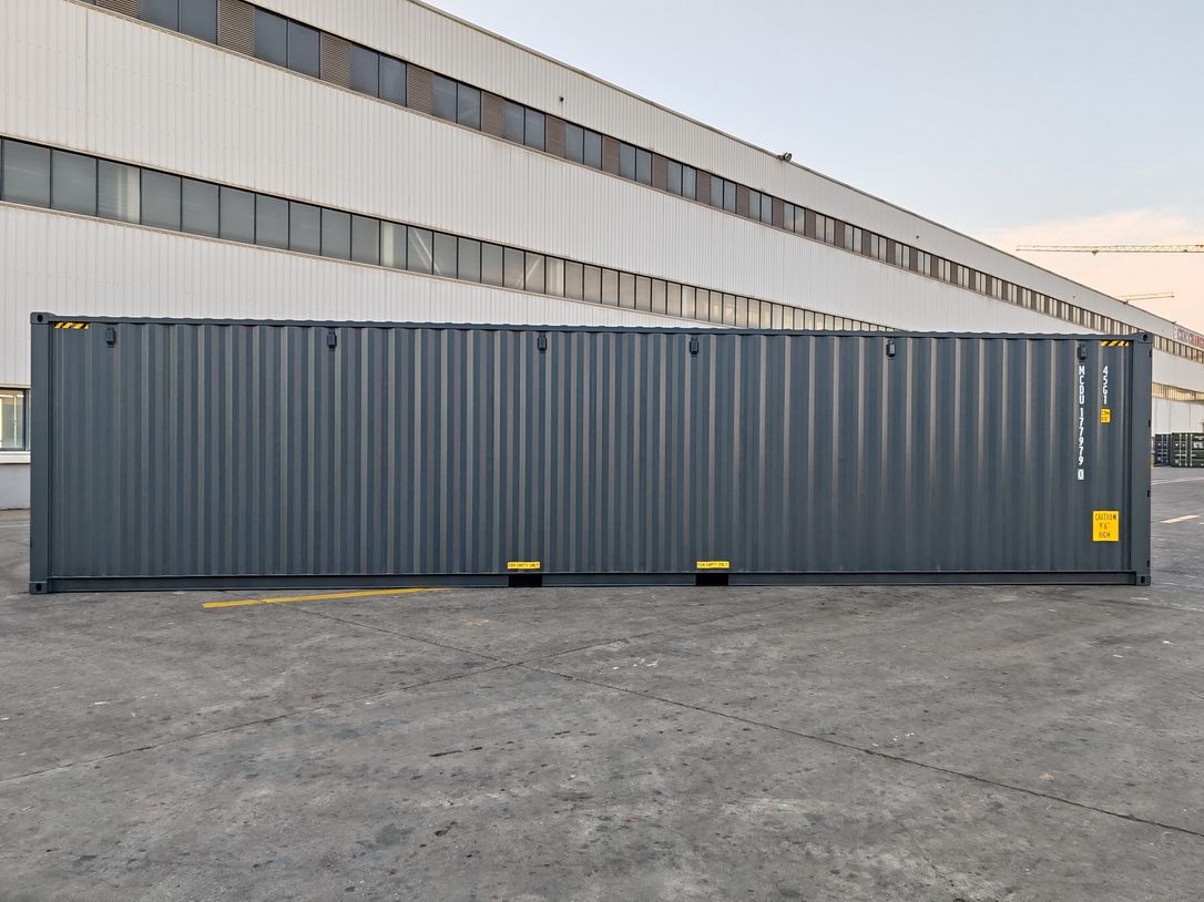 Container maritime 40 pieds High Cube gris anthracite vu de profil sur une aire de stockage, paroi ondulée et marquages d'origine, bâtiment industriel à l'arrière-plan