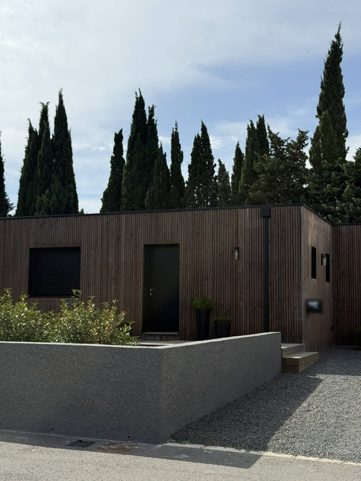 Maison container de plain-pied à Villedubert, près de Carcassonne : bardage bois à claire-voie vertical, toiture terrasse et menuiseries anthracite, derrière un muret bordé de cyprès