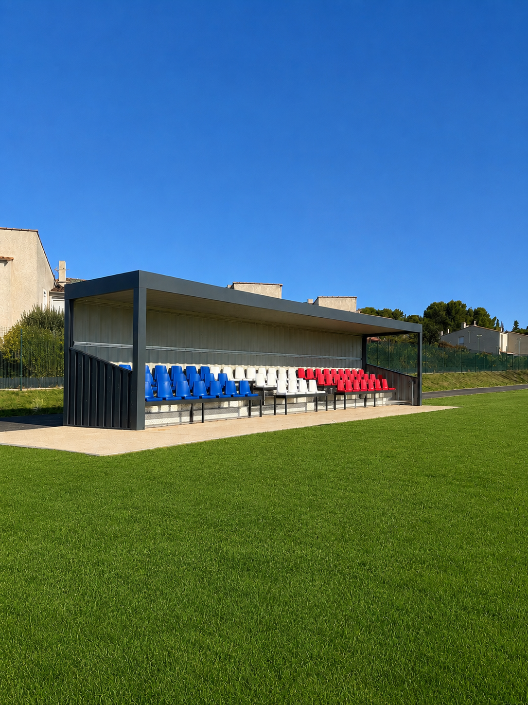 Tribune simple en container de 61 places au bord d'un terrain engazonné, toit plat anthracite et sièges bleus, blancs et rouges